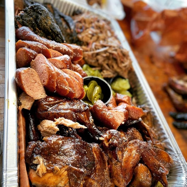 A large catering tray from Smith Spot BBQ filled with smoked sausage, pulled pork, chicken, and pickles.