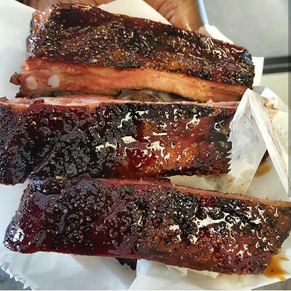 Three glistening Smith Spot BBQ ribs, coated in dark sauce and spices, resting on white paper.