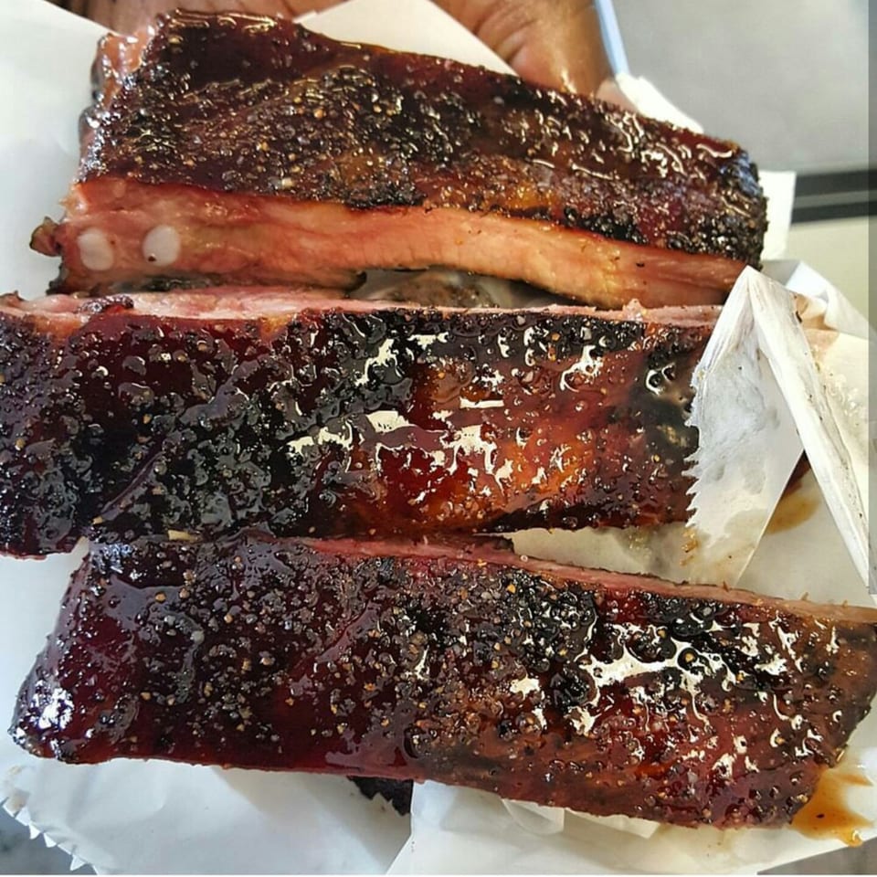 Three glistening Smith Spot BBQ ribs, coated in dark sauce and spices, resting on white paper.