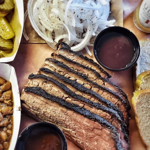 Slices of Smith Spot BBQ brisket with sides of pickles, onions, baked beans, and sauce.